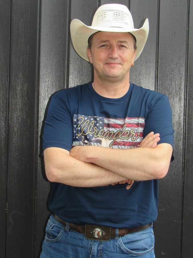 Patje Stew met gekruiste armen en witte cowboyhoed tegen een zwarte houten wand